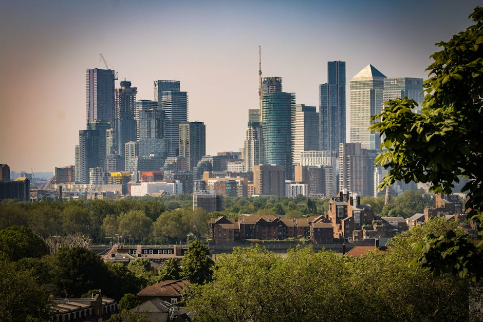 A cityscape view of London's skyline with multiple tall skyscrapers and modern office buildings, including several with glass facades reflecting sunlight, set against a pale blue sky. In the foregroun