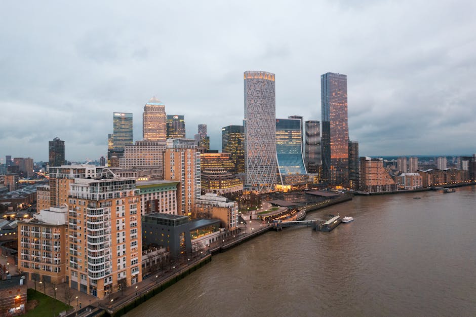 A cityscape view of London's skyline with multiple tall skyscrapers and modern office buildings, including several with glass facades reflecting sunlight, set against a pale blue sky. In the foregroun