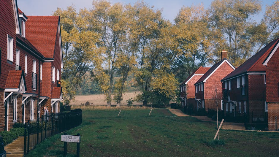A residential garden area featuring a gently sloping lawn with freshly trimmed grass, bordered by brick terraced houses with pitched roofs and small front yards. The garden bed along the fence contain